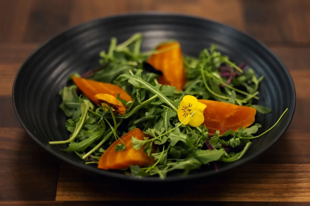Arugula and golden beet salad with an edible pansy, plated in a black bowl on dark wood