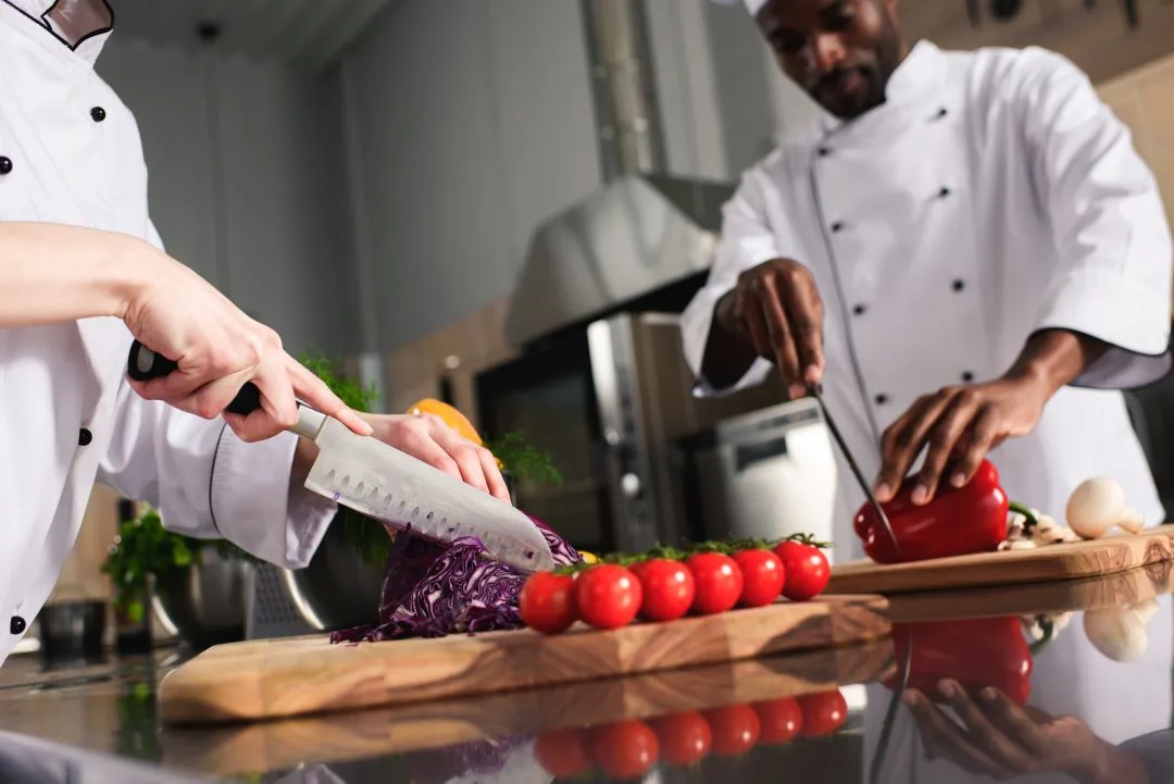Two chefs in white coats prepping cabbage, tomatoes and peppers on wooden boards in a stainless kitchen
