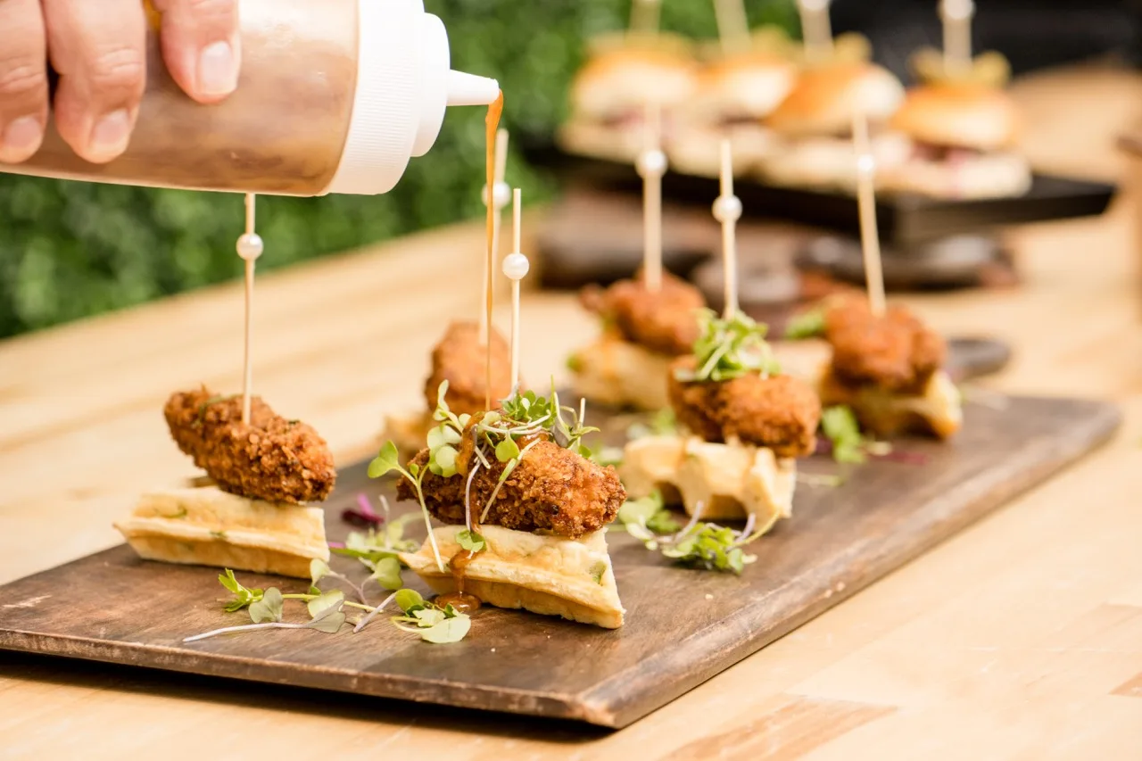 Chicken-and-waffle bites on skewers being drizzled with sauce on a wooden board, sliders behind