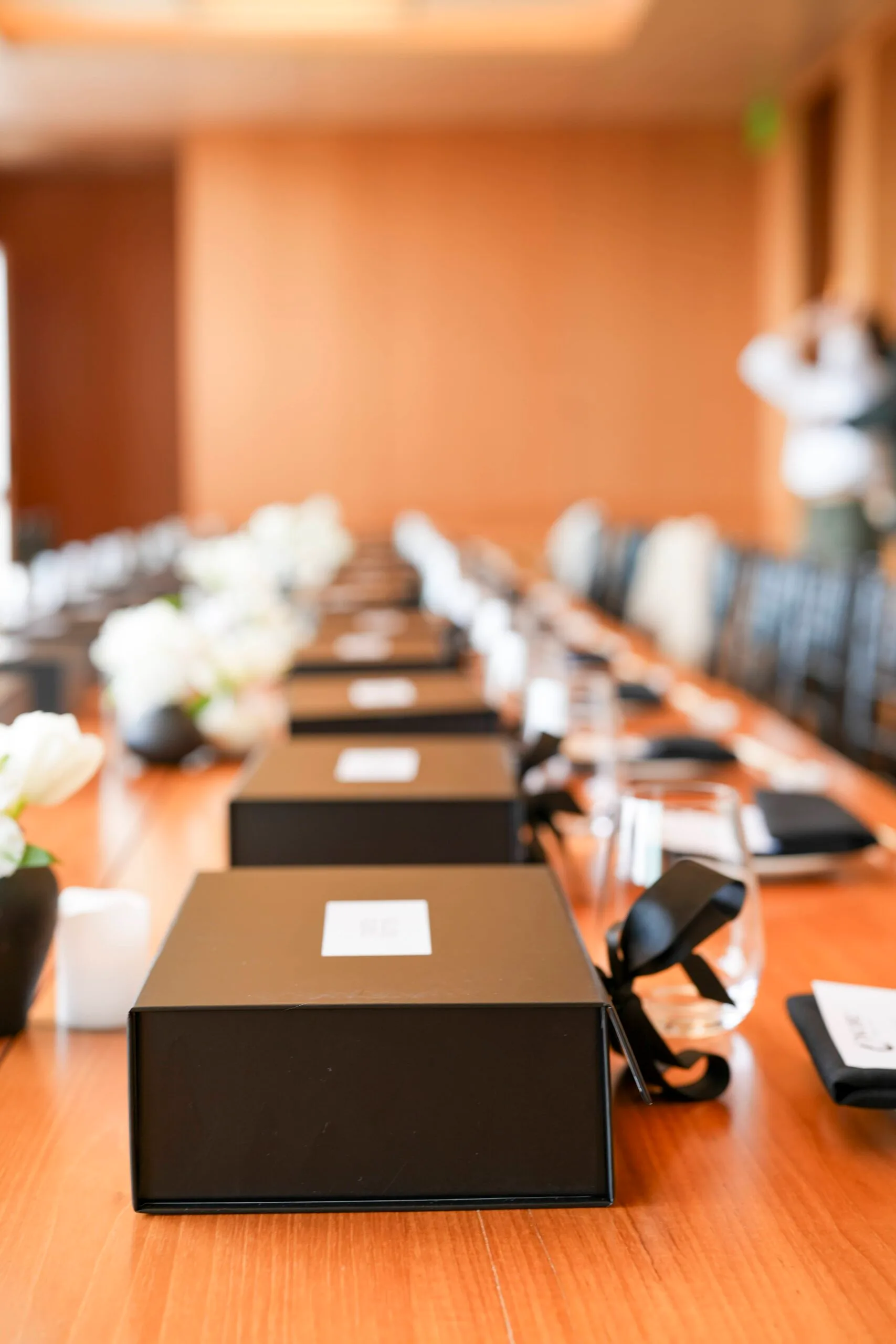 A row of ribbon-tied black meal boxes down a polished boardroom table set with white florals and glassware