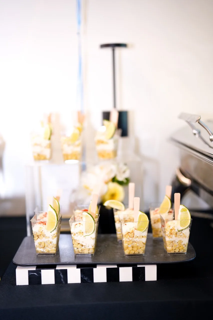 Individual street-corn cups with lime wedges and paddle picks on a black tray