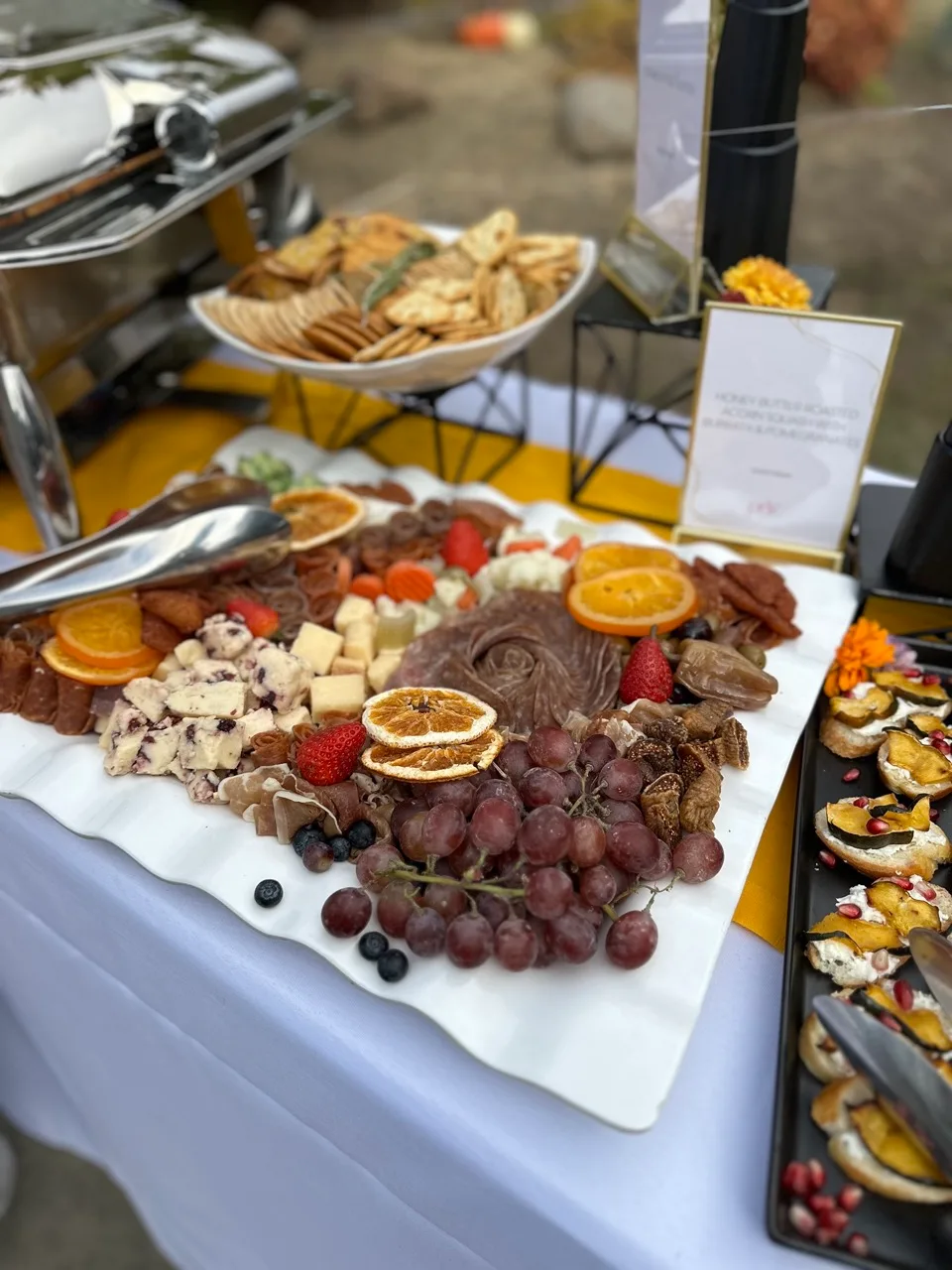Outdoor grazing table of cheeses, charcuterie, grapes, dried orange and squash crostini beside a chafing dish