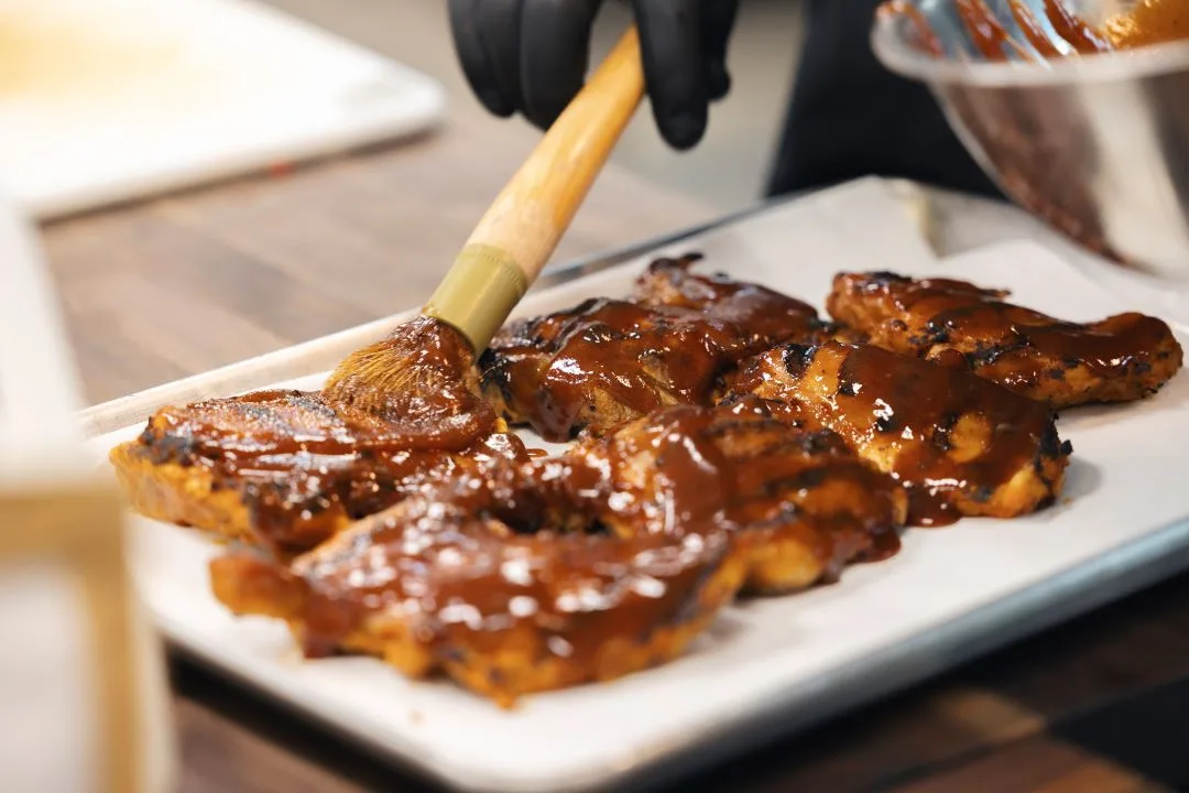 A gloved cook brushing barbecue glaze onto grilled chicken on a white platter