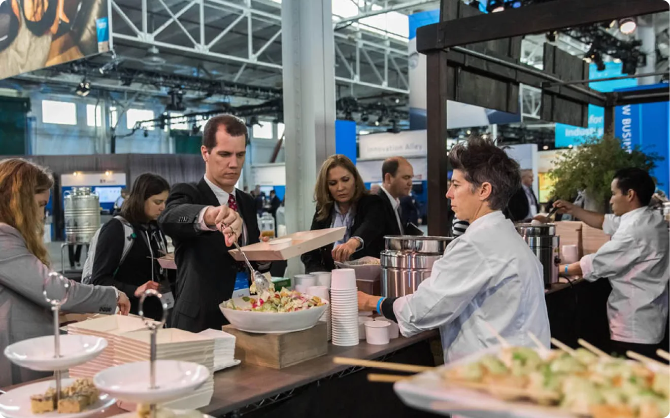 Attendees serving themselves from a staffed catering station on a conference floor, chefs plating behind the counter