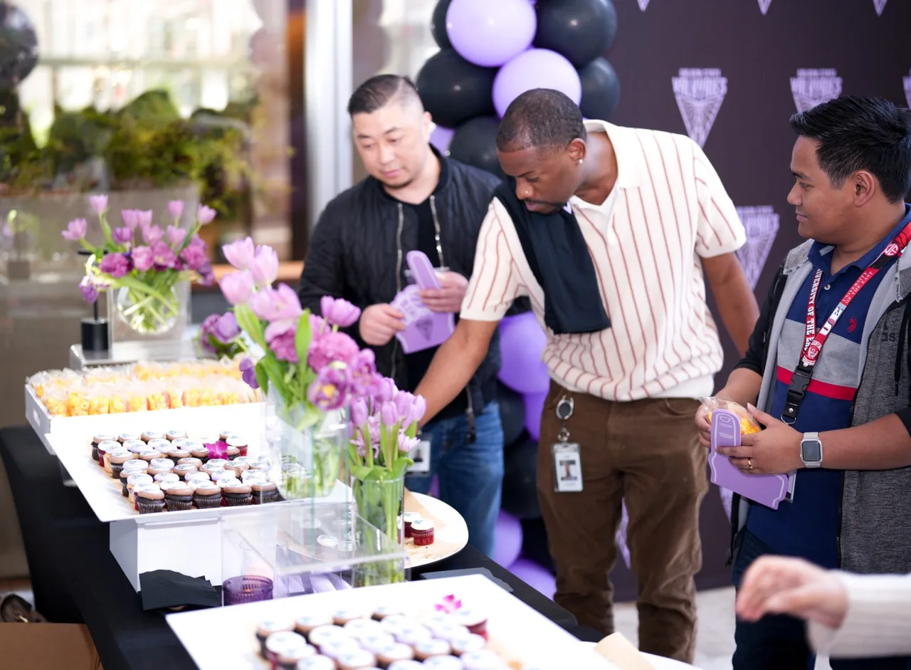 Office dessert table of logo-topped cupcakes with purple tulips and a balloon arch behind