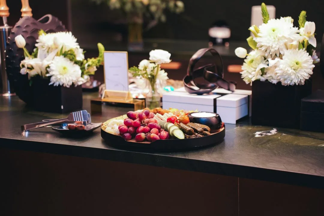 Grazing board of grapes, cheese and stuffed grape leaves on a dark stone counter, framed by white floral arrangements