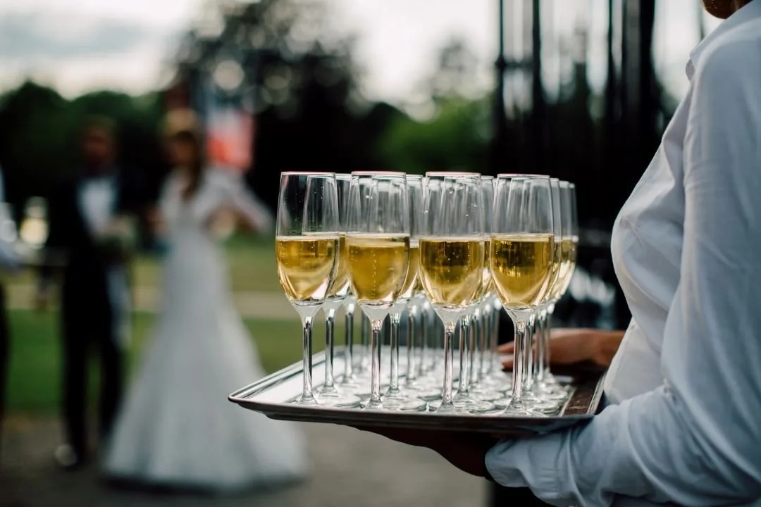 A server carrying a tray of champagne flutes at an outdoor wedding, the couple out of focus behind